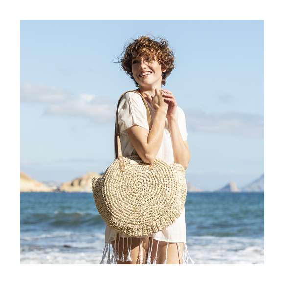 Bolso de desenfadado diseño en rafia sintética y con forma circular, un must. Compartimento principal de cierre de cremallera y tirador metálico, con bolsillo en el interior. Forro interior y asas largas en PU, con triple refuerzo en X y superficie para personalización en madera, especialmente diseñada para marcaje en láser.
