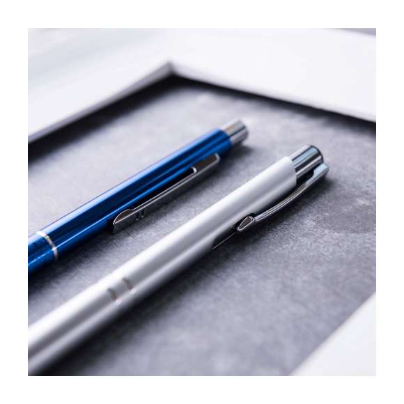 Original bolígrafo de mecanismo pulsador en elegante diseño bicolor. Con cuerpo troquelado en aluminio de suave acabado en variados colores. Original clip, anillas y punta en plateado. De cartucho jumbo y tinta azul.